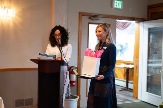 One woman speaks at podium while another woman stands to her right.