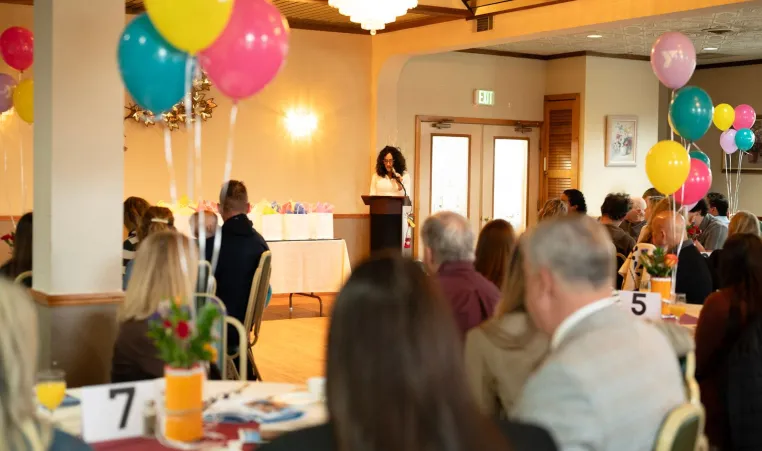 Woman standing at podium speaks to groups sitting at tables.