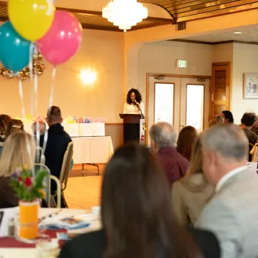 Woman standing at podium speaks to groups sitting at tables.
