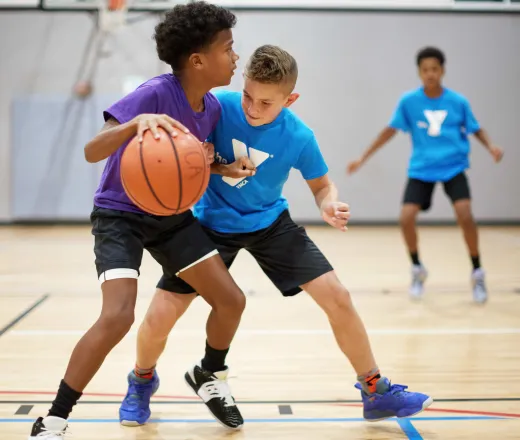 A boy attempts to dribble a basketball closer to the hoop while another teenage boy on the opposing team tries to block him from getting closer at an indoor basketball game.