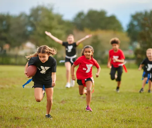 Five youth run down the flag football field during an outdoor game with one at the front carrying the football.